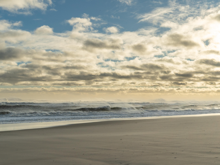 Waves crash on a sandy beach as the sun sets on a cloud-strewn sky.