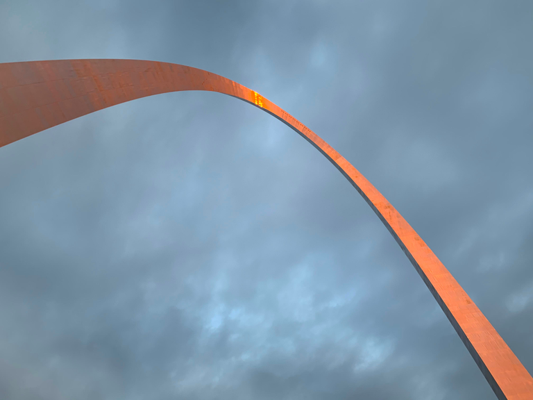 looking up at the Gateway Arch against a cloudy blue sky with a orange reflection at the top